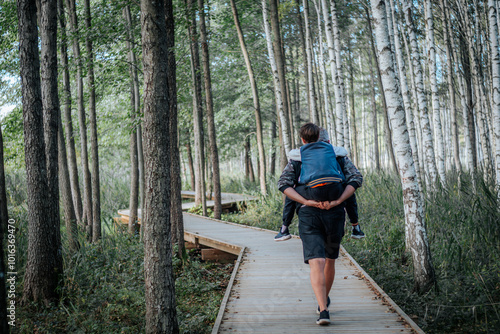 A smiling father carries his son in a piggyback while walking along a wooden path in the forest. The couple enjoys the outdoors surrounded by tall trees and nature.