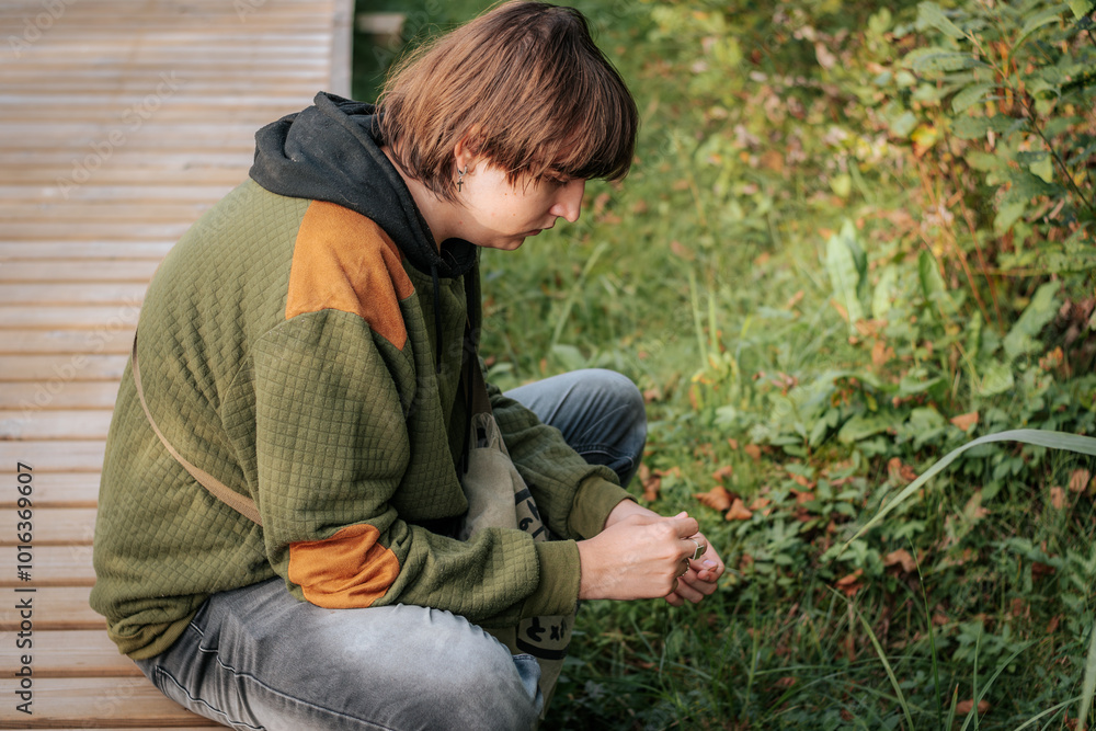 A teenage boy is sitting on the edge of a wooden path surrounded by greenery, wearing a green and orange jacket. He gazes thoughtfully into the distance, immersed in the natural environment. A teenage boy is sitting on the edge of a wooden path surrounded by greenery, wearing a green and orange jacket. He gazes thoughtfully into the distance, immersed in the natural environment.