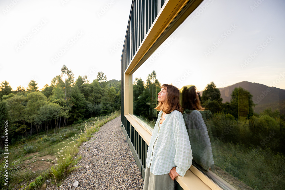 A woman leans against a modern cabin, reflecting on the tranquility of nature surrounding her. The calm of the mountains and the peace of solitude are captured in this serene moment