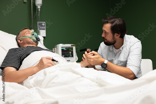 The older gentleman in the hospital bed wears an oxygen mask while his caring son sits vigilantly nearby, providing reassurance and encouragement. photo