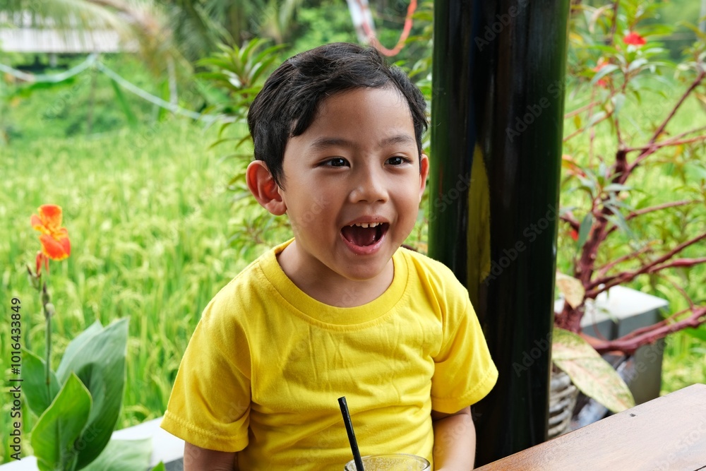Smiling boy in a yellow shirt enjoying a drink with a straw in a garden setting. The green background and colorful flowers create a vibrant, refreshing atmosphere, Smiling boy in a yellow shirt enjoying a drink with a straw in a garden setting. The green background and colorful flowers create a vibrant, refreshing atmosphere,