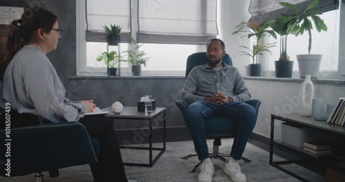 African American adult man talks to female therapist, discuses psychological trauma on therapy. Side view of psychologist sitting in chair with notebook, listening to patient, conducting consultation.