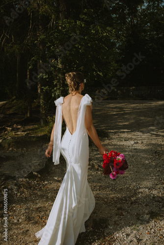 A modern bride in a white, open-back wedding dress walks along a gravel path, holding a vibrant bouquet of red and pink flowers, surrounded by natural woodland scenery.