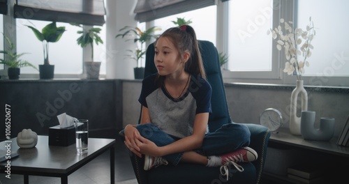 Asian kid sits in chair, talks to professional therapist during counseling session. Female child psychologist asks questions to school-aged girl, conducts therapy. Psychology and emotional support.