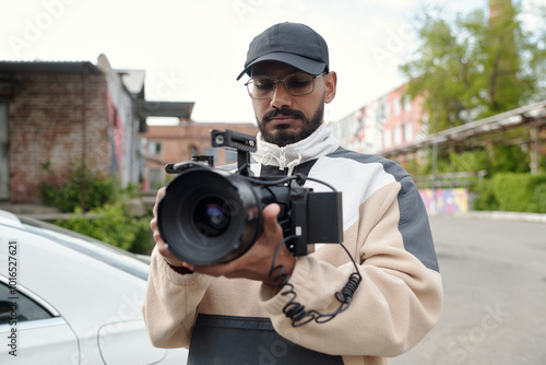 African American man wearing casual attire holding and adjusting large camera in outdoor urban area with buildings in background and parked cars visible