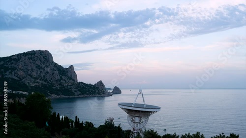Beautiful aerial view of the landscape of the sea and mountain with high fluffy pink pastel clouds and the radio telescope satellite dish, astrophysical antenna at sunset time
