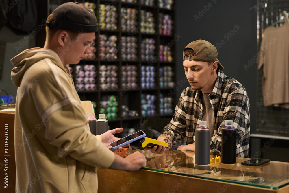 Young serious male shop assistant with payment terminal standing by counter in front of guy paying for graffiti paints Young serious male shop assistant with payment terminal standing by counter in front of guy paying for graffiti paints