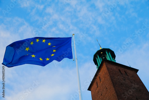 The European Union flag flutters in the breeze beside a tall brick tower of Stockholm City Hall, with a bright blue sky creating a vibrant backdrop during the day. photo