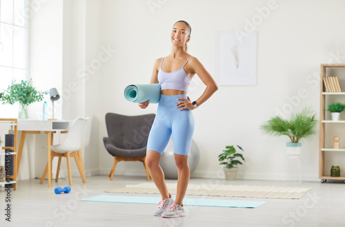 Portrait of a smiling athlete woman standing with a training mat at home. She is ready for workout or resting after sport session, embodying active fitness and sport in domestic gym.