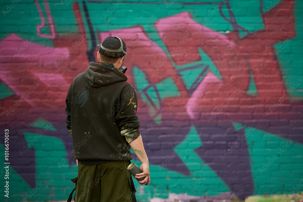 Back view of young man with dispenser containing graffiti paint standing in front of green wall with unfinished painting Back view of young man with dispenser containing graffiti paint standing in front of green wall with unfinished painting