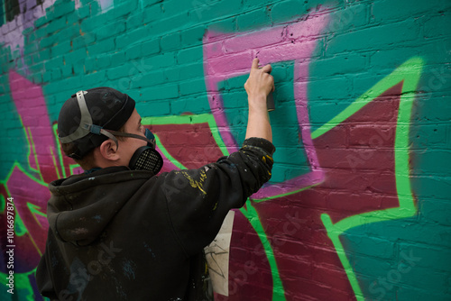 creative guy in respirator, black hoodie and beanie cap spraying pink paint from can on brick wall of building while creating artwork photo