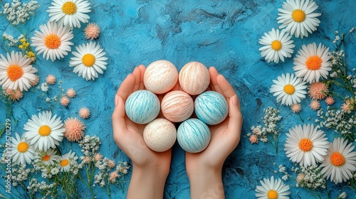 A pair of hands gently cradles six beautifully decorated Easter eggs, with pastel swirls. The background features a bright blue surface, adorned with fresh daisies and delicate flowers, creating a che photo