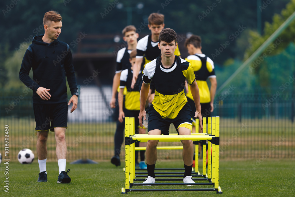 Obraz premium Teenagers on soccer training camp. Boys practice football with young coaches. Junior-level athletes improving soccer skills on outdoor training. The player jumping over hurdles and ladder-skipping