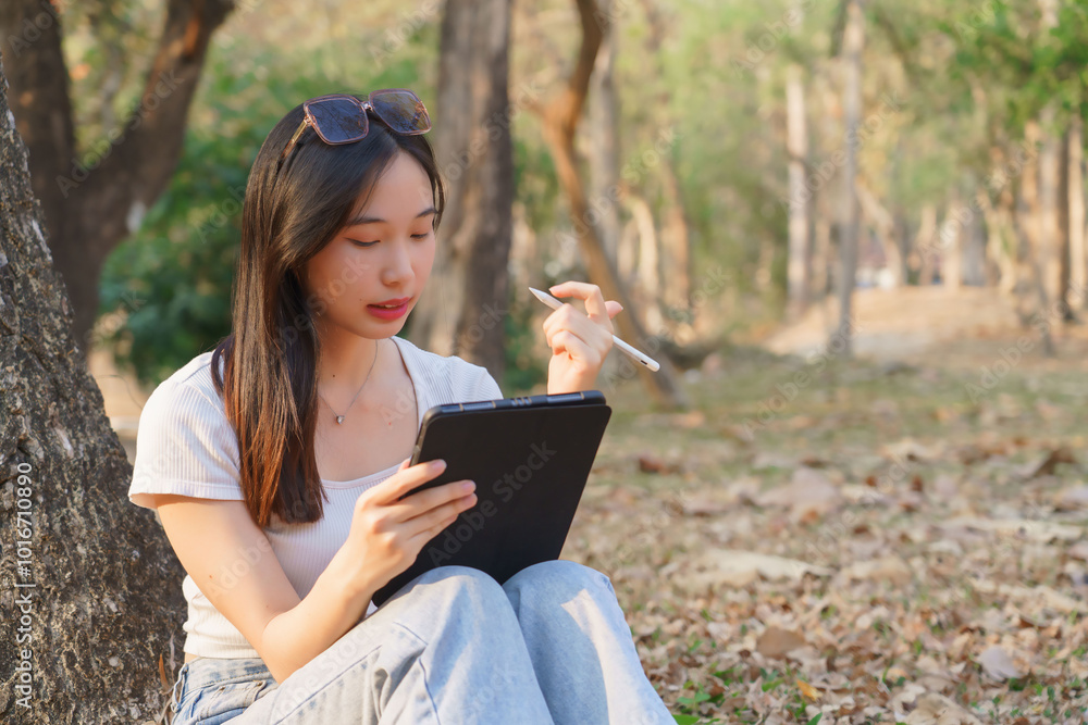 Young asian women writing business data and journey diary on digital tablet while sitting on the grass under tree in the nature park to working outside and relaxation with journey travel lifestyle