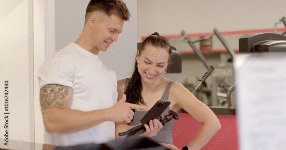 They look at the screen and analyze the actions. In a modern gym, a personal trainer is helping a woman with her workout plan using a tablet for assistance They look at the screen and analyze the actions. In a modern gym, a personal trainer is helping a woman with her workout plan using a tablet for assistance