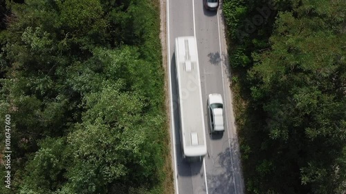 A view of the two-lane road that goes through the forest. A gray car and white car passes a white bus moving in the opposite direction. Aerial view directly above