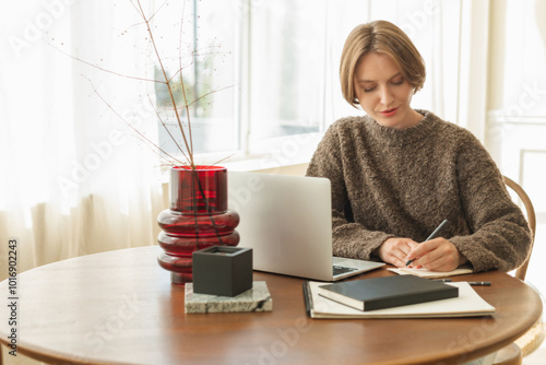 Young woman sitting at her dining table using her laptop and writing in a note pad photo