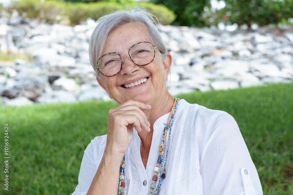 Portrait of attractive smiling senior woman of 70 years old sitting in the park looking at camera. Elderly grandmother with gray hair and eyeglasses relaxing enjoying a sunny day in outdoors Portrait of attractive smiling senior woman of 70 years old sitting in the park looking at camera. Elderly grandmother with gray hair and eyeglasses relaxing enjoying a sunny day in outdoors