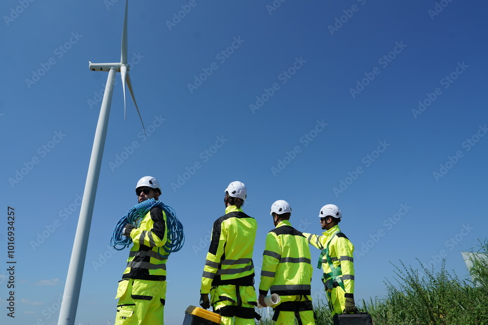 Four male engineers in uniforms standing on wind turbine station holding blueprints discussing and inspecting construction of wind turbine installation to generate electricity in wind farm. Clean ener Four male engineers in uniforms standing on wind turbine station holding blueprints discussing and inspecting construction of wind turbine installation to generate electricity in wind farm. Clean ener