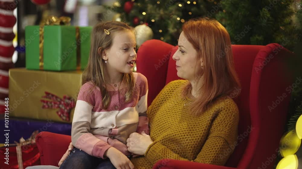 Medium shot of a young mother and her cute little daughter gossiping while sitting on sofa in the background of Christmas arrangement Medium shot of a young mother and her cute little daughter gossiping while sitting on sofa in the background of Christmas arrangement