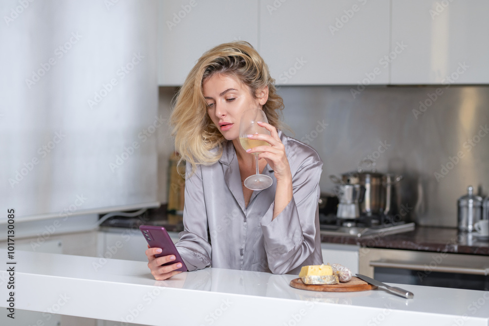Woman holding a glass of wine in her kitchen. Blond beautiful girl with phone drinking wine. Morning of pretty woman using social media on phone. Woman holding a glass of wine in her kitchen. Blond beautiful girl with phone drinking wine. Morning of pretty woman using social media on phone.