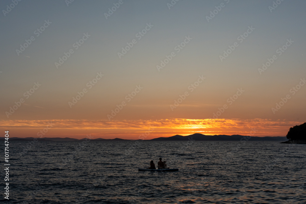 Fototapeta premium Family enjoying sunset paddleboarding on calm sea against a backdrop of hills and glowing sky. Concept of peaceful outdoor adventure, relaxation, and nature appreciation. High quality photo