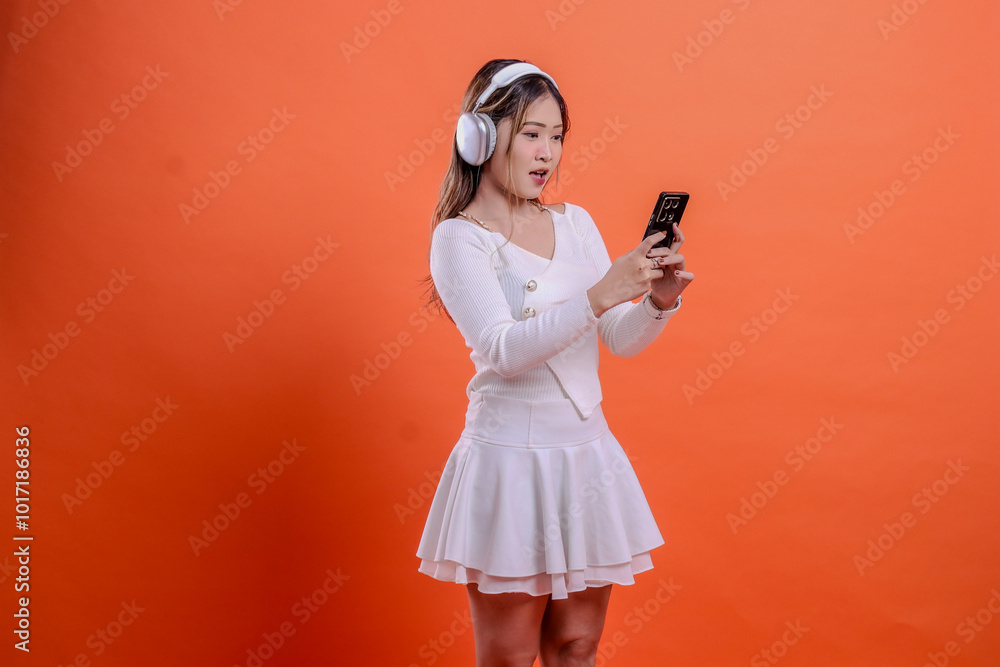 gesture young asian woman white top surprised to gadget wearing earphone headphone while holding mobile phone, cellphone, standing on orange background. for lifestyle, business, service concept gesture young asian woman white top surprised to gadget wearing earphone headphone while holding mobile phone, cellphone, standing on orange background. for lifestyle, business, service concept