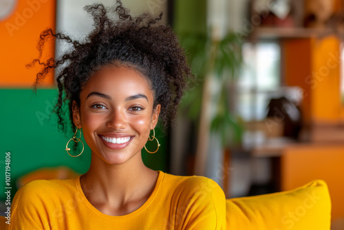 A cheerful, curly-haired woman smiles brightly indoors, wearing a vibrant yellow top. The colorful background adds warmth and liveliness to the scene. photo