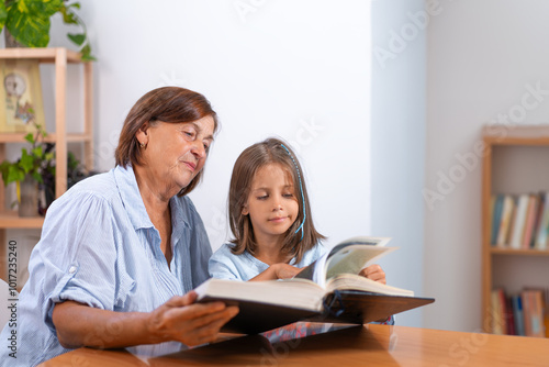 Grandmother and granddaughter reading book together at home. Cute girl turning pages as elderly woman sits with her, children education concept 