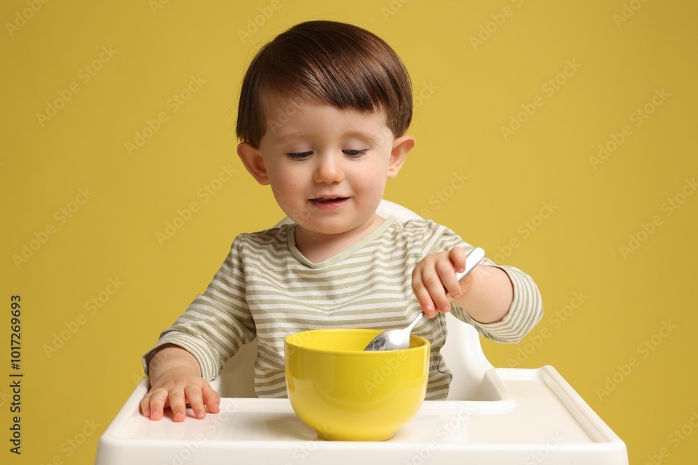 Cute little kid eating healthy baby food from bowl in high chair on yellow background Cute little kid eating healthy baby food from bowl in high chair on yellow background