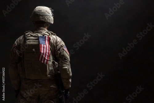 A soldier in full combat gear, stands with his back to the camera, with an American flag hanging from his tactical vest.