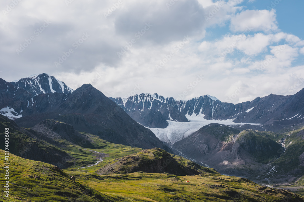 Dramatic scenery in alpine valley with creek among green hills and rocks with view to rocky pointy peak, large snow-capped peaked top, mountain range and big glacier tongue under clouds in blue sky. Dramatic scenery in alpine valley with creek among green hills and rocks with view to rocky pointy peak, large snow-capped peaked top, mountain range and big glacier tongue under clouds in blue sky.