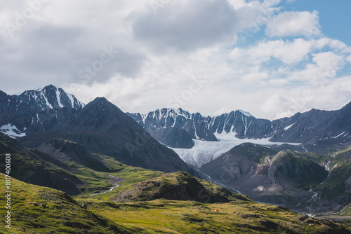 Dramatic scenery in alpine valley with creek among green hills and rocks with view to rocky pointy peak, large snow-capped peaked top, mountain range and big glacier tongue under clouds in blue sky.