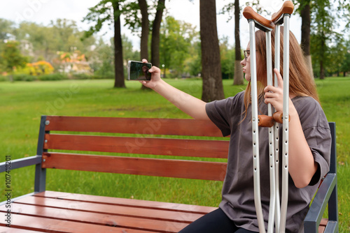 Obraz na plátně Teenage girl with a leg cast seated on a park bench, capturing a bittersweet selfie as a memento of her injury and the time lost during the recovery process