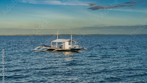 A beautiful traditional Filipino double-outrigger dugout bangka boat in the blue ocean. On the deck there are seats for tourists, a canopy, wooden masts. Ripples on the water. Clouds in the azure sky. photo