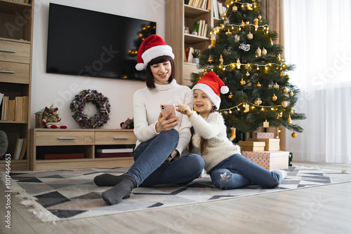 Caucasian mother and young daughter wear Santa hats, sit on floor in cozy room, use smartphone together by Christmas tree. Warm family moment captures holiday joy, bonding, and festive spirit.