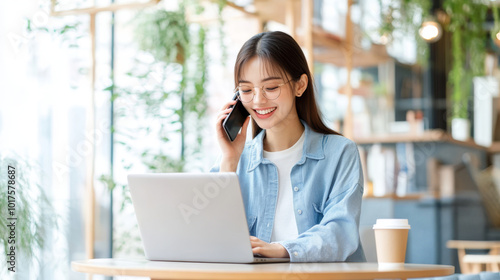 A young woman is engaged in phone conversation while working on her laptop in cozy cafe filled with greenery. Her cheerful demeanor reflects productive and pleasant atmosphere