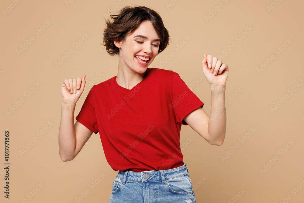 Young smiling happy woman she wear red t-shirt casual clothes doing winner gesture celebrate clenching fists dance isolated on plain pastel light beige background studio portrait. Lifestyle concept. Young smiling happy woman she wear red t-shirt casual clothes doing winner gesture celebrate clenching fists dance isolated on plain pastel light beige background studio portrait. Lifestyle concept.