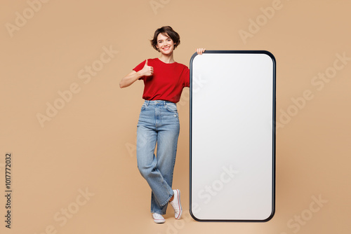 Full body young woman she wear red t-shirt casual clothes big huge blank screen mobile cell phone smartphone with area show thumb up isolated on plain pastel light beige background. Lifestyle concept. photo