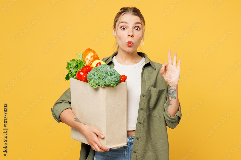 Young shocked surprised woman wear casual clothes hold bag for takeaway mock up with food products show ok okay gesture isolated on plain yellow background. Delivery service from shop or restaurant. Young shocked surprised woman wear casual clothes hold bag for takeaway mock up with food products show ok okay gesture isolated on plain yellow background. Delivery service from shop or restaurant.