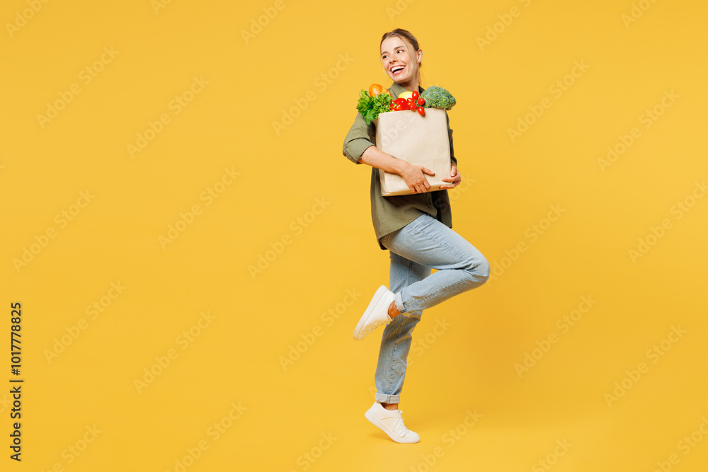 Full body side view young smiling happy woman wear casual clothes hold bag takeaway mock up with food products look aside raise up leg isolated on plain yellow background. Delivery service from shop. Full body side view young smiling happy woman wear casual clothes hold bag takeaway mock up with food products look aside raise up leg isolated on plain yellow background. Delivery service from shop.