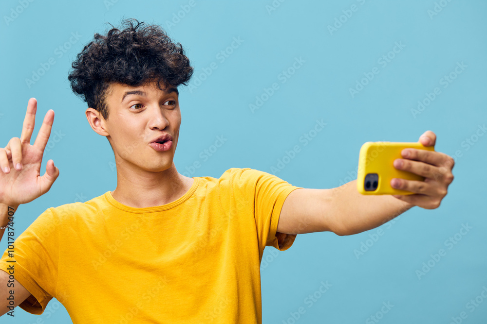 Young man taking a selfie with a smartphone, cheerful expression, wearing a yellow shirt, against a light blue background, perfect for social media and lifestyle concepts Young man taking a selfie with a smartphone, cheerful expression, wearing a yellow shirt, against a light blue background, perfect for social media and lifestyle concepts
