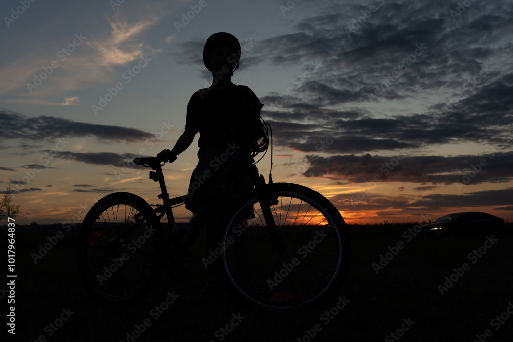 Fototapeta premium Silhouette of a cyclist standing beside their bike at sunset, capturing the peacefulness of dusk in the countryside. High quality photo