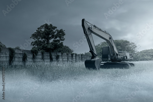 Large construction machine is sitting in field with concrete wall and trees. The sky is cloudy and dark.