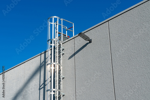 A sturdy metal ladder extends from the ground to the rooftop of a building, reaching towards a clear blue sky without any clouds, highlighting a sunny day and the urban architecture.