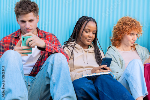 A diverse group of three smiling young students sit together on the floor, smiling and interacting on their smartphones against a blue background. photo