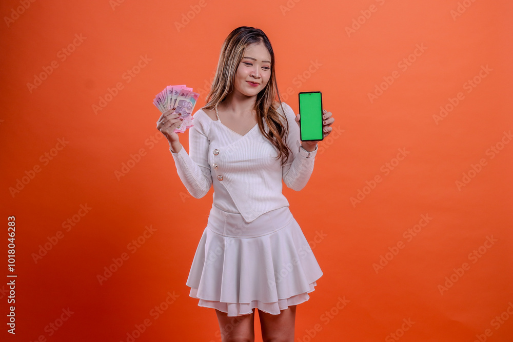 charm young beautiful asian woman white top standing smile look at gadget hand hold rupiah cash money and cellphone screen, smartphone isolated orange background. for service concept, business charm young beautiful asian woman white top standing smile look at gadget hand hold rupiah cash money and cellphone screen, smartphone isolated orange background. for service concept, business