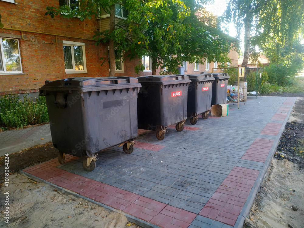 Rows of black waste bins lined neatly along a newly paved walkway in a residential area during the late afternoon Rows of black waste bins lined neatly along a newly paved walkway in a residential area during the late afternoon
