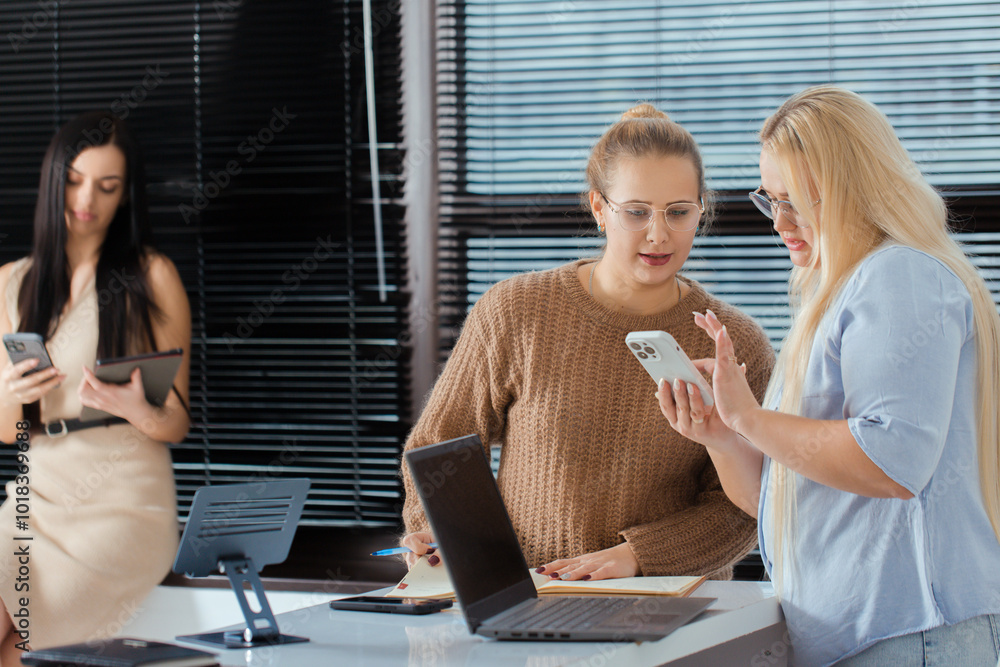 Two women stand at the table and discuss something while looking at the phone, the third woman looks at the tablet by the window