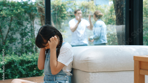 Stressed and unhappy young girl huddle in corner, cover her ears blocking sound of her parent arguing in background. Domestic violence at home and traumatic childhood develop to depression. Synchronos